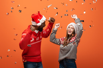 happy interracial couple in santa hats and Christmas sweaters showing yes gestures under confetti...