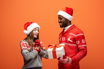african american man gifting Christmas present to happy girlfriend on orange background