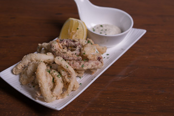 Simple white plate with fresh fried calamari with dipping sauce and slices of lemon, isolated over wooden background.