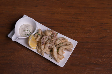 Simple white plate with fresh fried calamari with dipping sauce and slices of lemon, isolated over wooden background.