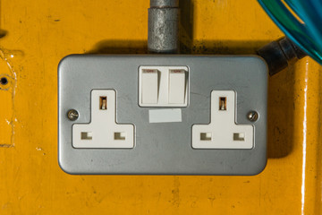 Power outlets on a bright yellow orange metal beam in an industrial factory settings with colourful blue pipes in the corner of the image.