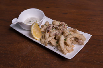 Simple white plate with fresh fried calamari with dipping sauce and slices of lemon, isolated over wooden background.
