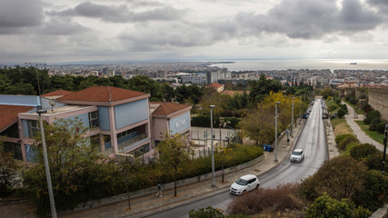 A view of Thessaloniki from atop the old town on a moody partly cloudy day. Cars travelling down the road with the new area to the left and the old town to the right overlooking the city scape