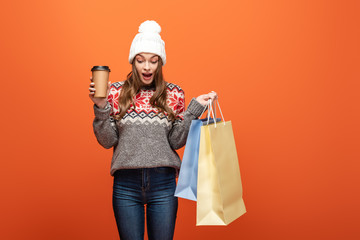 shocked girl in winter outfit holding shopping bags and coffee to go on orange background