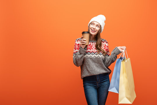 happy girl in winter outfit holding shopping bags and coffee to go on orange background