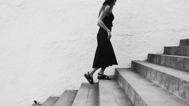 Girl with long hair, black dress and black hat walking up the stairs against a white wall. Sunny day. Concrete stair. Elegant young woman in black hat. Fashion.