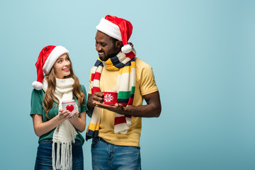 Fototapeta premium happy interracial couple in santa hats and scarves holding mugs with cocoa isolated on blue