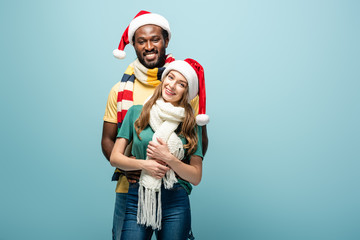 Fototapeta premium happy interracial couple in santa hats and scarves hugging isolated on blue