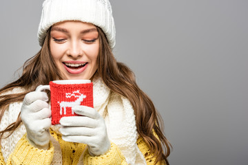 happy girl in yellow sweater, scarf, hat and gloves holding mug of cocoa isolated on grey