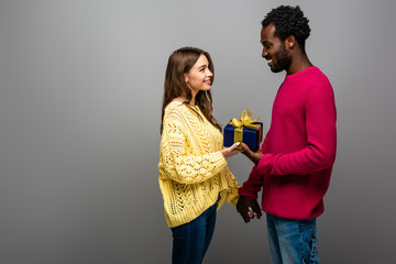 happy interracial couple in sweaters holding present on grey background
