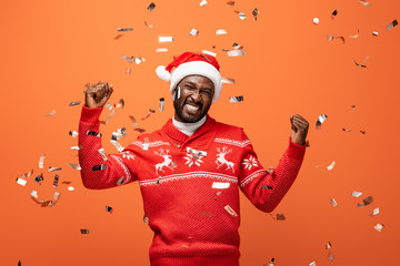 happy man in santa hat under falling confetti showing yes gesture on orange background