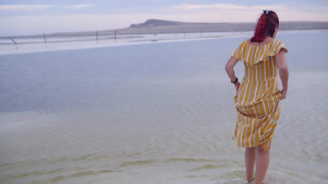 Young thin girl in yellow striped dress comes slowly into water of salt lake in shoes . The expanse of the Bascunchak salt lake at sunset during summer holidays. Bogdo hill