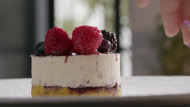 A close up of a spinning appetizing dessert with a thick layer of white souffle and a handful of different berries on the top. A hand puts beside a tiny flower twig