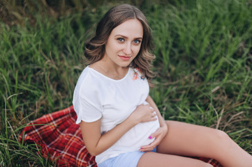 Naklejka na ściany i meble Portrait of a pretty woman expecting a baby in nature. A young and pregnant blonde girl in a white dress sits on a plaid in green grass and holds on to her stomach. Pregnancy picture, concept.
