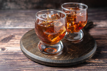Two glasses of rum or bourbon with ice on rustic wooden background.