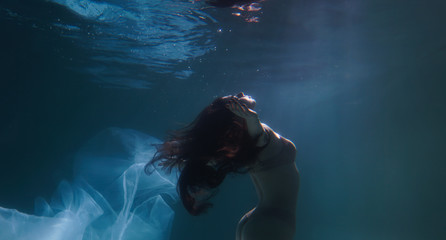 Obraz na płótnie Canvas Beautiful girl swims underwater with long hair. Blue or gold background like gold. The atmosphere of a fairy tale or magic. Diving under the water with a shiny cloth