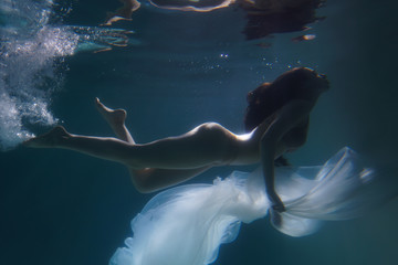 Beautiful girl swims underwater with long hair. Blue or gold background like gold. The atmosphere of a fairy tale or magic. Diving under the water with a shiny cloth