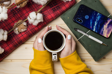 hands holding a cup of coffee on a wooden background, on which lie branches of cotton and a red tablecloth, top view