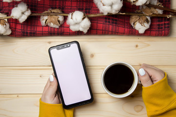 hands holding a cup of coffee on a wooden background, on which lie branches of cotton and a red tablecloth, top view