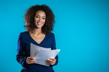 Pretty african american female boss checks documents, utility bills. Serious businesswoman standing on blue background. She is satisfied with work of staff. 