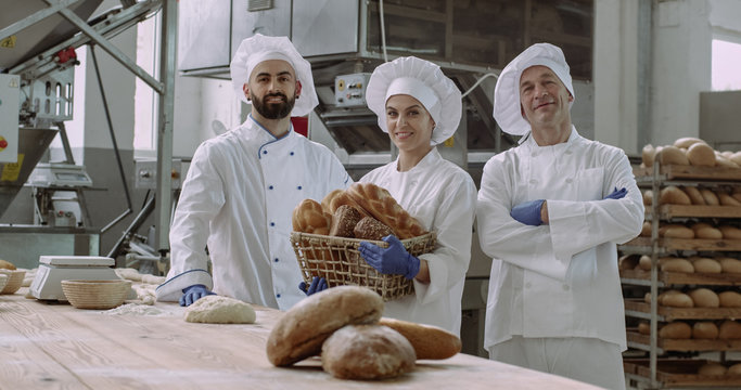 Group of three commercial faces of bakers holding a vintage basket with organic bread looking straight of the camera wearing special uniform smiling pretty big bakery industry factory food