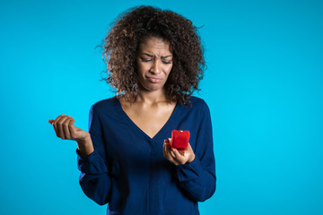 Pretty african-american young woman holding small jewelry box on blue background. Disappointed...