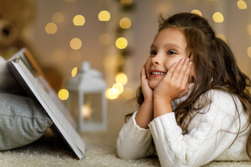 Happy smiling little girl is reading fairy tale book on the background with lights. child dreams of...