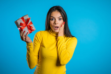 Beautiful surprised spanish woman received gift box with bow. She is happy and flattered by attention. Girl on blue background. 