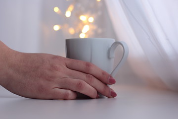 Woman is holding a white porcelain cup with fairy lights in the background. 
