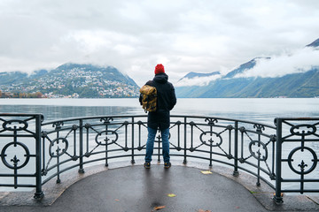 Man with backpack enjoy panorama on pier in Lugano. Man in travel. Lake Lugano, southern slope of Alps. Landscape in Switzerland. Amazing scenic outdoors view. Canton of Ticino. Adventure lifestyle