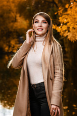 A beautiful blonde in a beige coat walks in the autumn in the park. Yellow leaves, autumn, nature. Autumn photo shoot in the forest