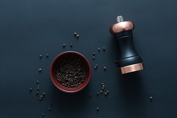 Pepper mill with copper inserts on black background with some black pepper in a clay bowl.