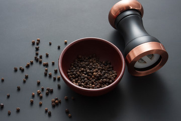 Pepper mill with copper inserts on black background with some black pepper in a clay bowl.