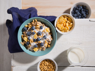 Bowl with oatmeal, corn flakes, blueberries and a glass of milk on a white wooden background