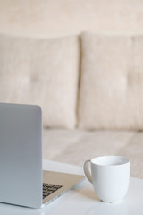 Gray laptop with a white mug on the table in the room. Work at home. Close up. Vertical.