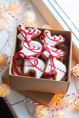 Traditional handmade deer shaped gingerbread cookies decorated with icing and ribbons in the carton box on the windowsill, the Christmas light garlands around