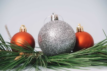 Close up of a big silver glitter christmas ornament next to two smaller red mate christmas balls on top of pine needles on a white background
