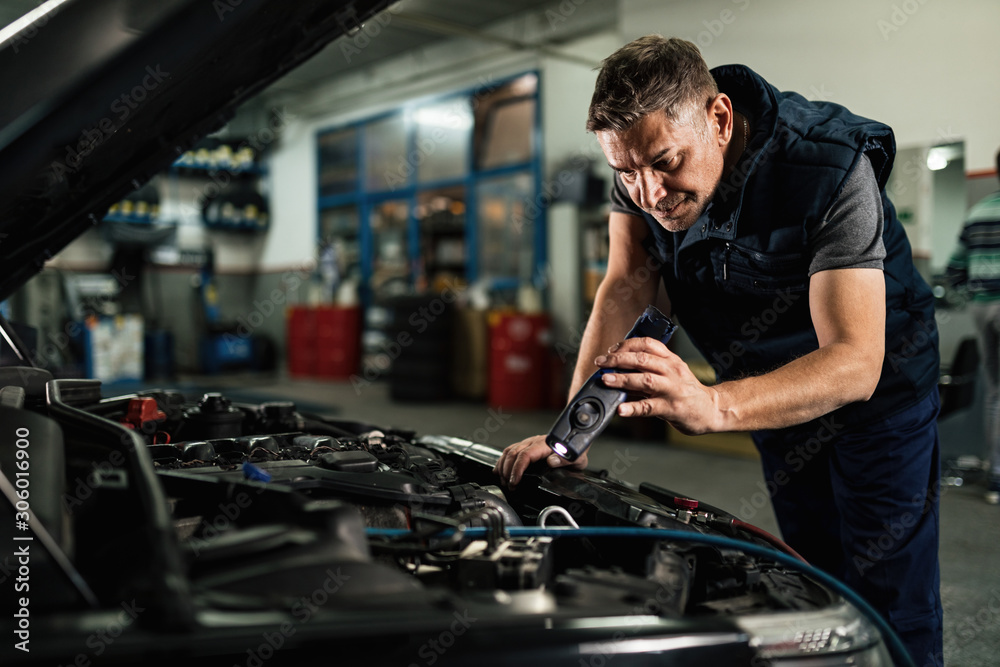 Wall mural auto mechanic using lap while examining car engine in repair shop.