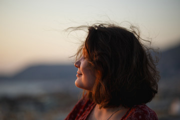 Summer outdoor side portrait of young pretty sensual girl posing at roof at sunset. Fluing hair on wind. View on mediterranian city buildings and red roofs.