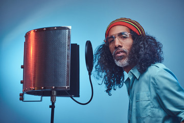 African Rastafarian vocal artist wearing a blue shirt and beanie standing next to a microphone...