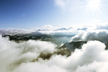 Photos of snowy Swiss mountains, seen from the top of the Stanserhorn. Sun in the center that illuminates the valley, with clouds passing between the mountains, creating many colors.