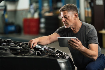 Car mechanic using digital tablet while examining engine in auto repair shop.