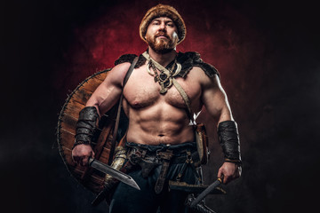 Serious Viking clad in light armor with a shield behind his back holds a sword and an axe. Posing on a dark background with red light