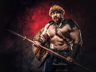 Fototapeta na wymiar Serious Viking clad in light armor with a shield behind his back holds a spear. Posing on a dark background with red light