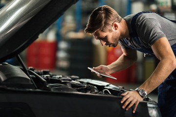 Young auto repairman examining car engine while using digital tablet in a workshop.