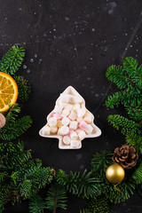 Marshmallows in Christmas tree shape plate on dark background with fir tree green branches. Smores for coffee or hot chocolate, copy space