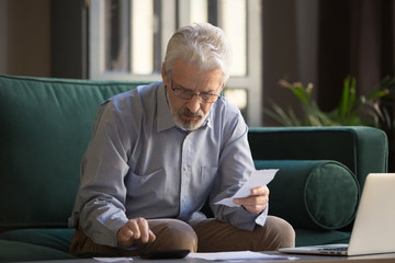 Mature man holding receipt calculates on calculator monthly expenses
