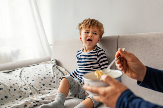Unhappy child refusing to eat at home with white background while father is trying to deceive the toddler by tricks. Upset unhappy child refuse to eat healthy food 