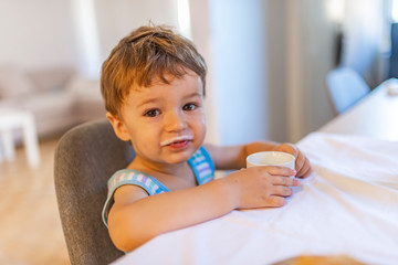 Toddler sitting in chair and eating greek yogurt. Baby learning to eat and has yogurt on face. Kid boy eating healthy food at home or daycare. Cute little boy eating yogurt.