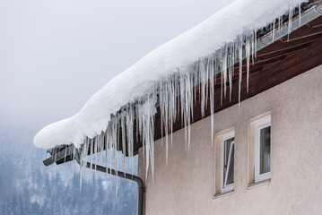 Big icicles and snow hanging over the rain gutter on a roof of a traditional wooden house in the mountains in winter could be dangerous. Blue sky at the background. - obrazy, fototapety, plakaty